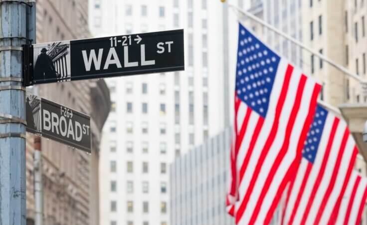 Street signs for Wall Street and Broad Street in New York City stand in front of two large American flags hanging from the side of a skyscraper. By kasto/stock.adobe.com