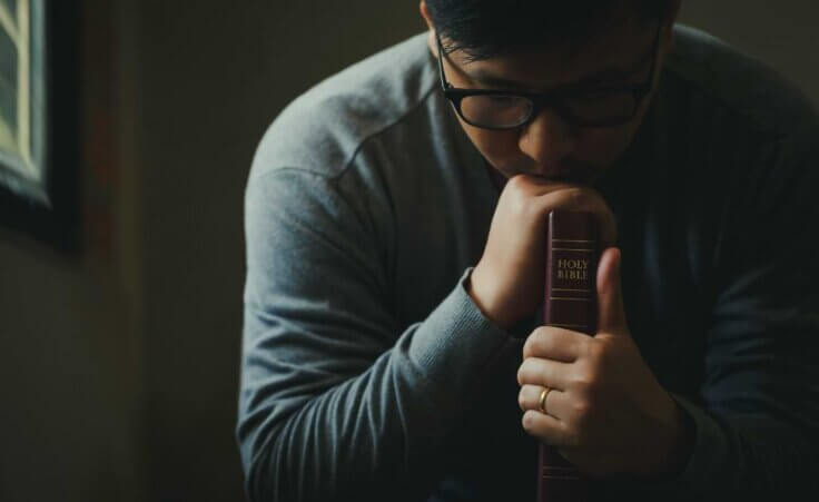 A man in glasses bows his head in prayer while holding a Bible upright on a table, its spine and the word "BIBLE" facing out. By Pcess609/stock.adobe.com. Many pastors and church leaders suffer from compassion fatigue.