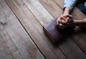 A man clasps his hands in prayer on top of a Bible sitting atop a wood table. By 4Max/stock.adobe.com | What does 1 Thessalonians 5 18 mean