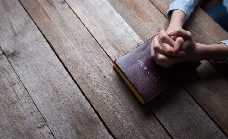 A man clasps his hands in prayer on top of a Bible sitting atop a wood table. By 4Max/stock.adobe.com | What does 1 Thessalonians 5 18 mean