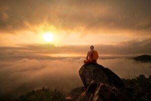 A Buddhist monk in orange robes sits atop a high mountain staring across an expanse into a rising sun. By ittipol/stock.adobe.com