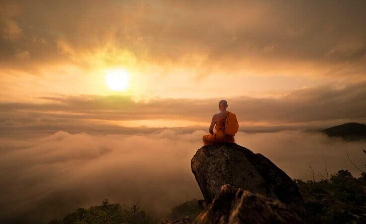 A Buddhist monk in orange robes sits atop a high mountain staring across an expanse into a rising sun. By ittipol/stock.adobe.com