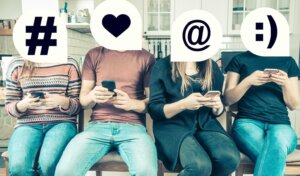 A group of four Gen Z young adults sit next to each other, each one looking at their phone, and each person's head is covered by an emoji. By rzoze19/stock.adobe.com