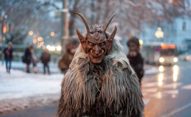 Outside at dusk on a snowy street, a person in a Krampus costume looks directly at the camera. Krampus is a "demonic anti-Santa" with goat horns. By Manfred Herrmann/stock.adobe.com