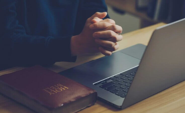 A man folds his hands in prayer above an open laptop on a table. A closed Bible sits to the right of the laptop. Pcess609/stock.adobe.com