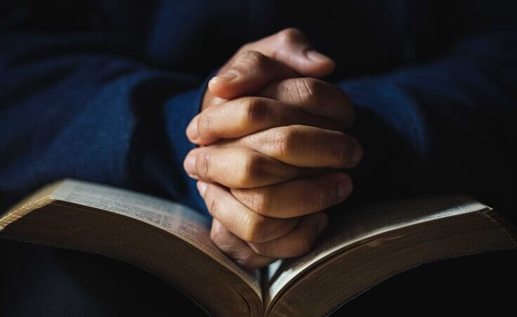 Hands folded in prayer on top of an open Bible, illustrative of spiritual enlightenment. By doidam10/stock.adobe.com