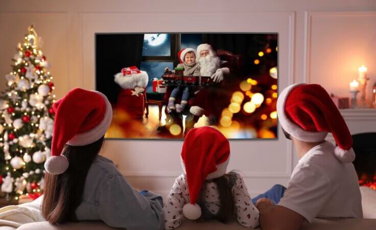 A family of three, all wearing Santa hats, sits on a couch while watching a Christmas movie featuring Santa Claus. By New Africa/stock.adobe.com