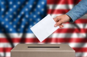 An extended arm drops a ballot into a ballot box in front of an American flag, a symbol of democracy. Why is democracy popular? By andriano_cz/stock.adobe.com