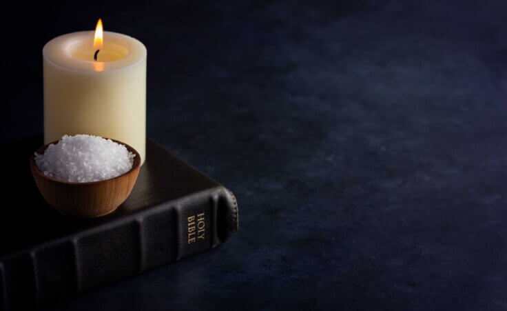 A lit candle and a handful of salt in a small bowl sit atop a black Bible. By pamela_d_mcadams/stock.adobe.com
