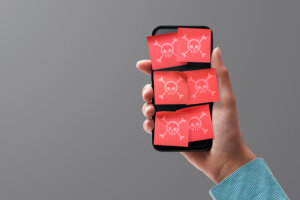 A lone hand holds up a smartphone covered in six red sticky notes, each of which has a poison symbol on it depicting malware. By stokkete/stock.adobe.com
