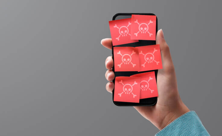 A lone hand holds up a smartphone covered in six red sticky notes, each of which has a poison symbol on it depicting malware. By stokkete/stock.adobe.com