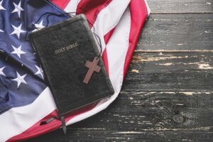 A small brown cross sits atop a closed black Bible, which sits atop an American flag. By Pixel-Shot/stock.adobe.com. The documentary "God & Country" looks at the rise of Christian nationalism.