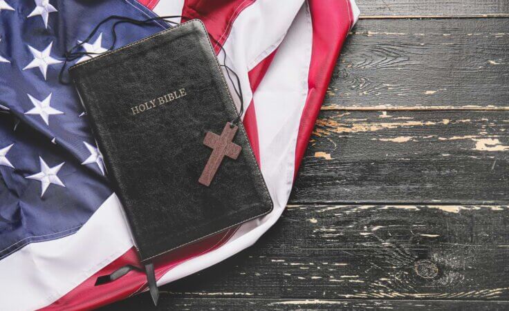 A small brown cross sits atop a closed black Bible, which sits atop an American flag. By Pixel-Shot/stock.adobe.com. The documentary "God & Country" looks at the rise of Christian nationalism.