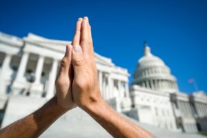 Hands folded in prayer in front of the US Capitol building. By lazyllama/stock.adobe.com