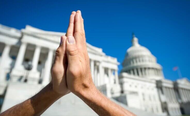 Hands folded in prayer in front of the US Capitol building. By lazyllama/stock.adobe.com