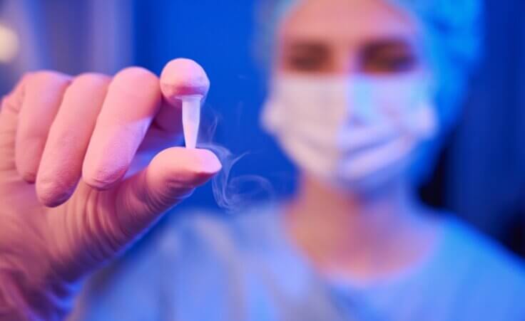 A lab scientist in a facemask holds up a vial containing frozen embryo samples. By Svitlana/stock.adobe.com