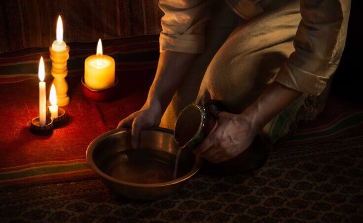 A stylized image of Jesus bending down and pouring water from a jar into a bowl in preparation to wash feet. The "He Gets Us" campaign, funded by Hobby Lobby's Green family, that aired during Super Bowl LVIII showed Christians washing others' feet. By Marina/stock.adobe.com