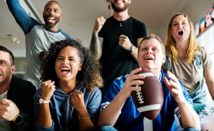 Three excited football fans sit on a couch while three more cheer behind them, a picture of a typical Super Bowl Sunday viewing party. By Rawpixel.com/stock.adobe.com