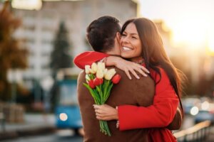 A woman in a red jacket hugs a man in a brown jacket. She's clutching a handful of flowers in celebration of Valentine's Day. By Sanja_85/stock.adobe.com