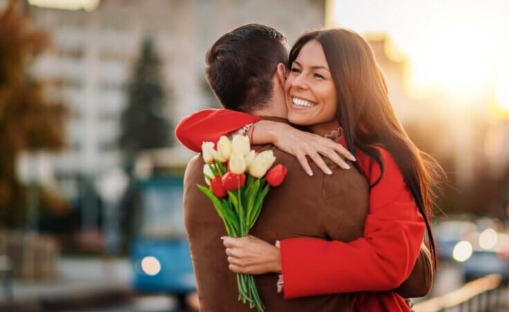 A woman in a red jacket hugs a man in a brown jacket. She's clutching a handful of flowers in celebration of Valentine's Day. By Sanja_85/stock.adobe.com