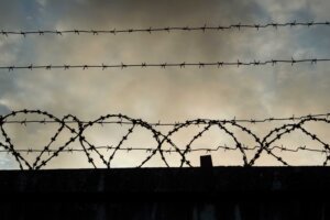 Barbed wire above a fence, reminiscent of Aushwitz, on a dark, cloudy day. By Buyan/stock.adobe.com