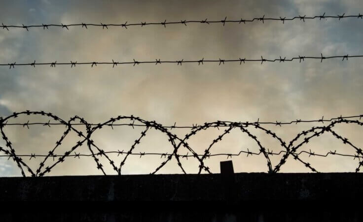 Barbed wire above a fence, reminiscent of Aushwitz, on a dark, cloudy day. By Buyan/stock.adobe.com