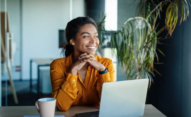 A woman at work with a beaming smile looks to the side in front of an open laptop and a mug, an illustration of happy people. By Prostock-studio/stock.adobe.com