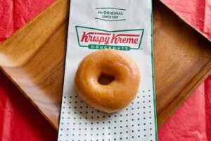 A single glazed donut sits atop a Krispy Kreme bag, which lays across a wooden plate on top of a red tablecloth. By EWY Media/stock.adobe.com