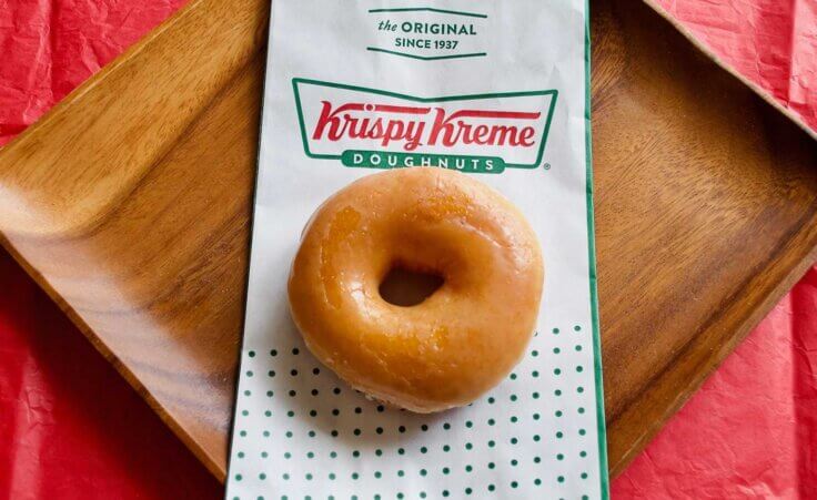 A single glazed donut sits atop a Krispy Kreme bag, which lays across a wooden plate on top of a red tablecloth. By EWY Media/stock.adobe.com