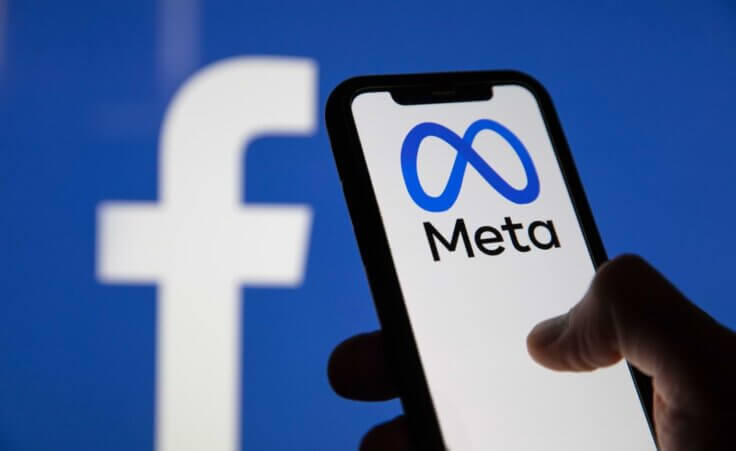 A hand holds a smartphone showing the Meta company logo in front of a screen showing the Facebook logo. Meta owns Facebook. By ink drop/stock.adobe.com