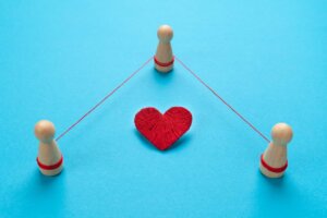 Three wooden figurines are tied together with red string around a woven heart, illustrating a throuple. By Andrii Zastrozhnov/stock.adobe.com