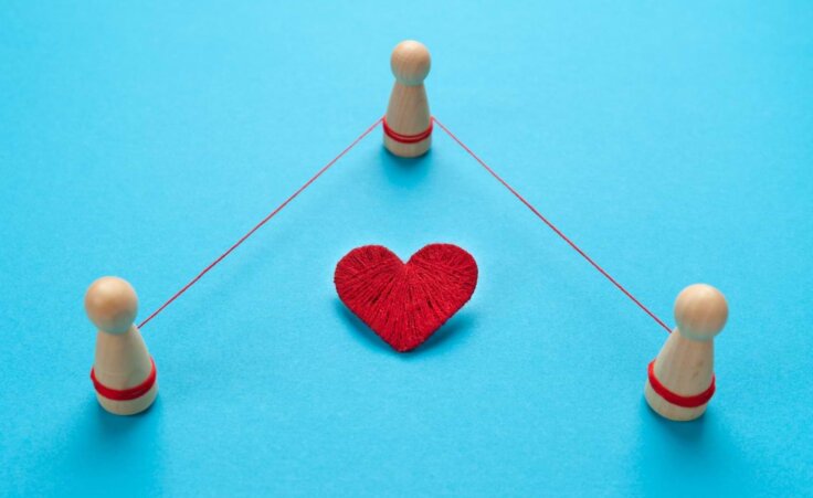 Three wooden figurines are tied together with red string around a woven heart, illustrating a throuple. By Andrii Zastrozhnov/stock.adobe.com