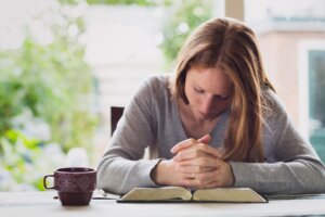 A woman bows her head and clasps her hands in prayer over an open Bible on a table with a cup of coffee nearby. By ptnphotof/stock.adobe.com