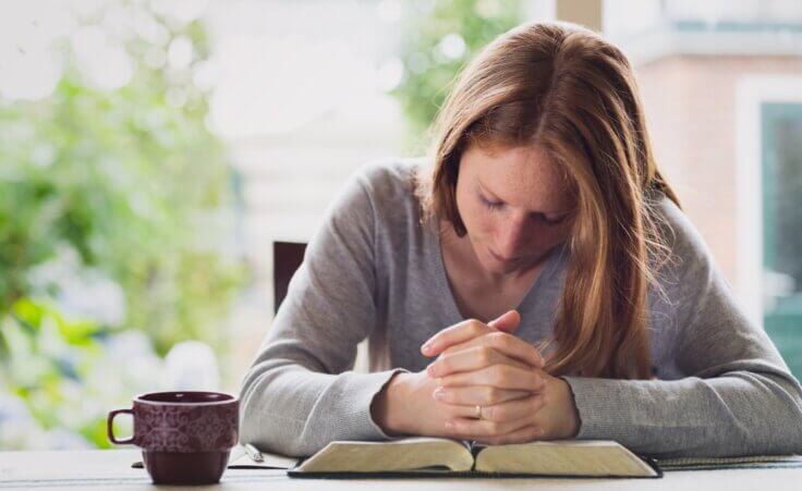 A woman bows her head and clasps her hands in prayer over an open Bible on a table with a cup of coffee nearby. By ptnphotof/stock.adobe.com