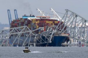 A boat moves past a container ship as it rests against wreckage of the Francis Scott Key Bridge on Tuesday, March 26, 2024, as seen from Pasadena, Md. (AP Photo/Mark Schiefelbein). The bridge disaster in Baltimore has resulted in at least 6 dead.