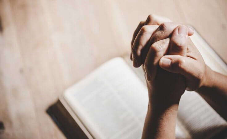 Hands folded in prayer over an open Bible. By doidam10/stock.adobe.com