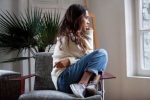 A young woman appears anxious and curled up while sitting in a chair and looking out of a window. By fizkes/stock.adobe.com