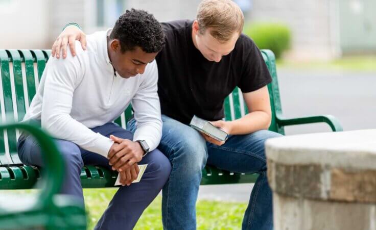 Two men bow their heads in prayer while sitting on a park bench. One man holds a closed Bible in his left hand while his right hand is on the other man's shoulder. By Lydia/stock.adobe.com