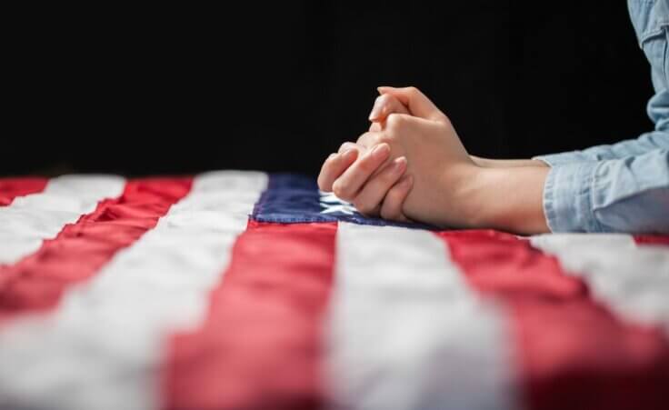 Clasped hands in prayer on top of an American flag. By 4Max/stock.adobe.com