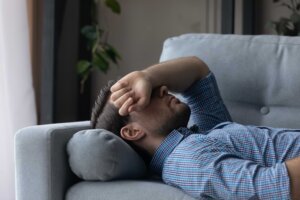 A man lies on his back on a couch, his arm covering his eyes, in a state of anxiety. By fizkes/stock.adobe.com