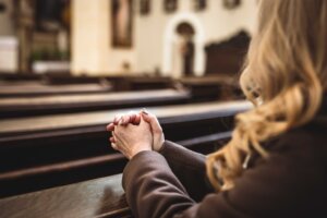 A woman sitting in a pew in an empty church clasps her hands in prayer. By encierro/stock.adobe.com