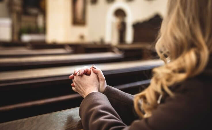 A woman sitting in a pew in an empty church clasps her hands in prayer. By encierro/stock.adobe.com