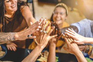 Adult friends in group, sitting around the dinner table high-fives all together celebrating friendship and to be united together - concept of lifestyle, trust, unity, togetherness - focus on hands. By Lomb/stock.adobe.com