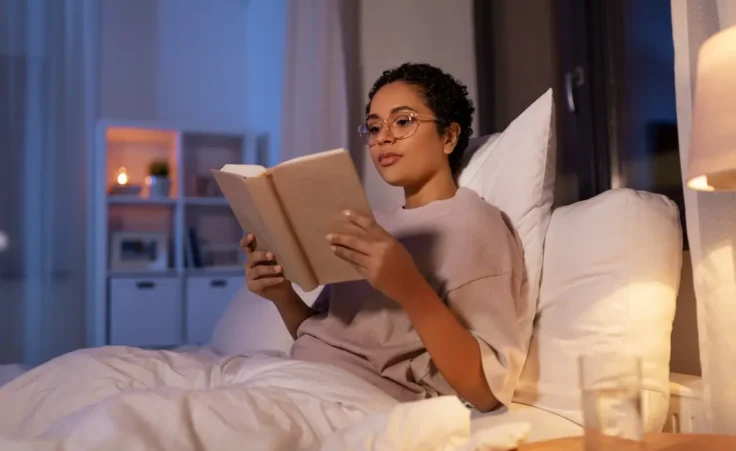 A woman reading a book in bed at home at night to illustrate healing through rest. By Syda Productions/stock.adobe.com