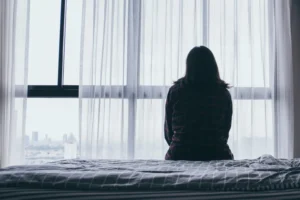 Woman sits on the edge of a bed and looks out the window on a dreary day. By panitan/stock.adobe.com. Jen Hatmaker