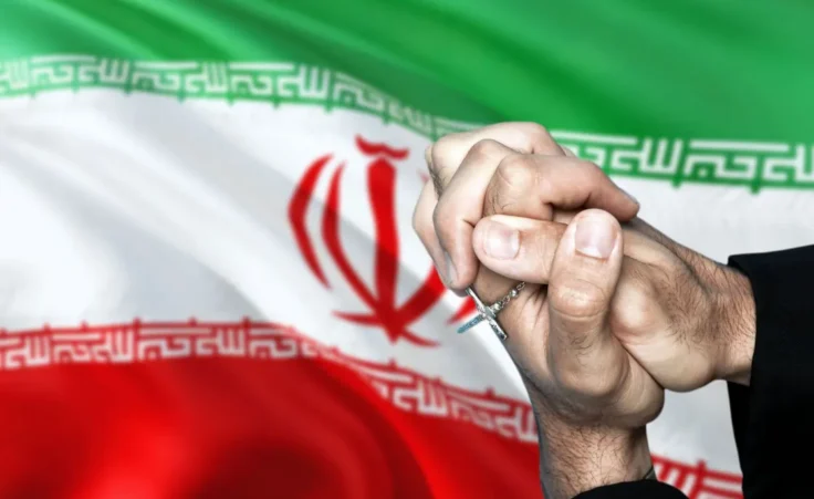 Iran flag and praying patriot man with crossed hands. Holding cross, hoping and wishing. By sezerozger/stock.adobe.com