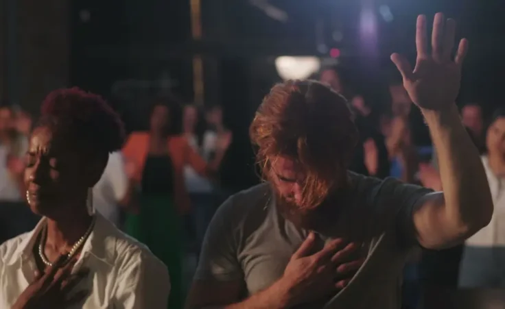 Congregation in prayer during spiritual gathering, hands on hearts, diverse group expressing faith and devotion, solemn worship moment with illuminated cross in background. By Marco/stock.adobe.com