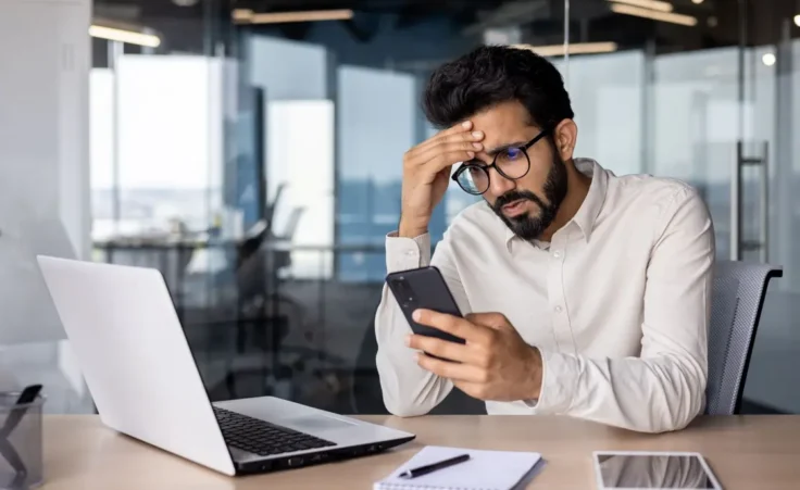 Upset and worried young Indian man sitting in the office at the desk, holding his head and looking frustrated at the phone screen By Tetiana/stock.adobe.com