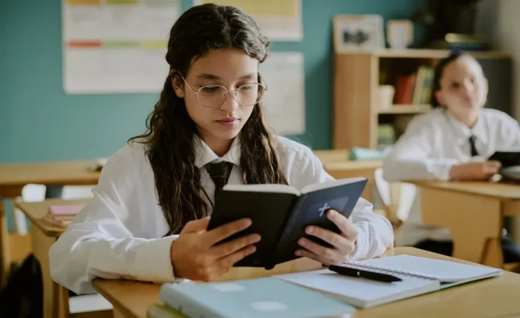 Student with glasses intensely reading Bible in classroom. Setting including open notebooks on desks and another student in background by AnnaStills/stock.adobe.com