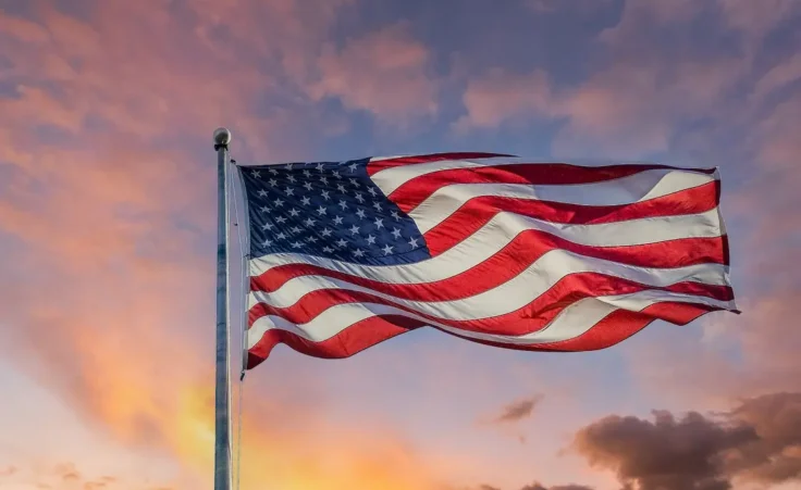 An american flag backlit by the sun against a blue sky with clouds by dbvirago/stock.adobe.com